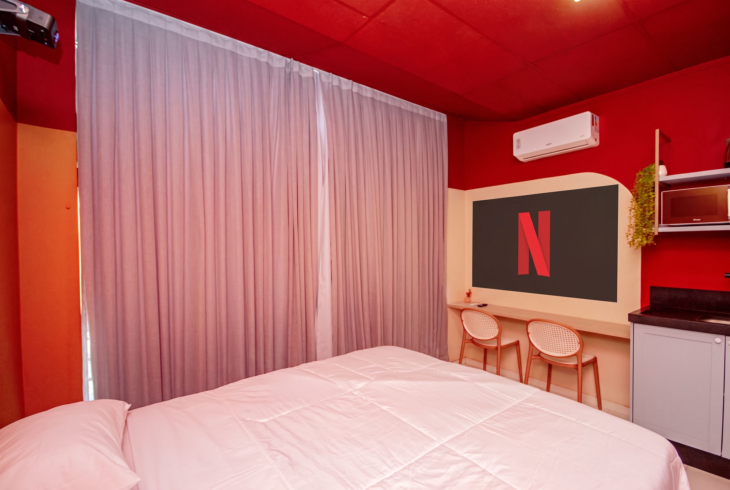 A hotel room with a bed, a desk with two chairs, a large window with curtains, an air conditioning unit, and a wall-mounted TV.