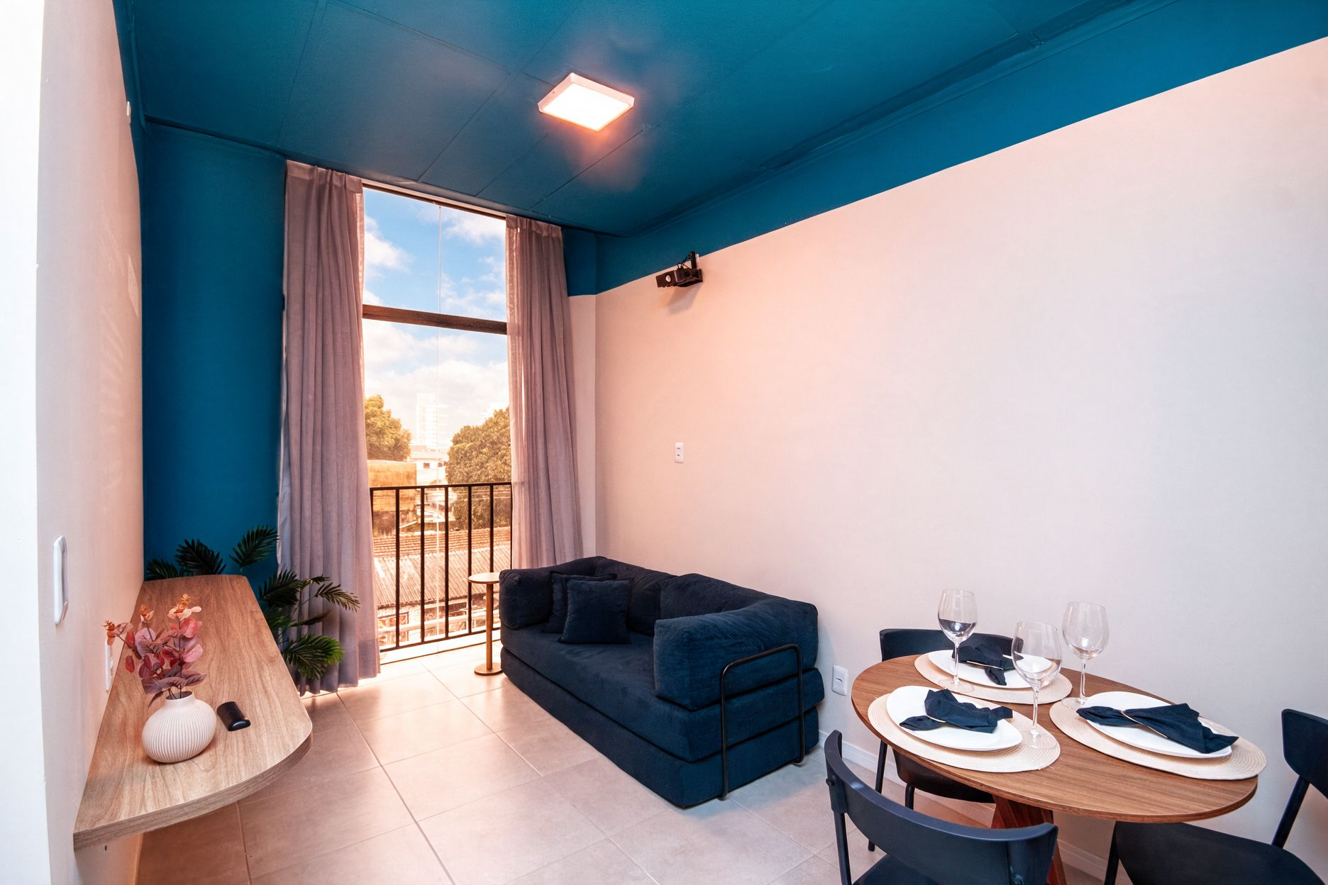 Uma sala de estar moderna com um sofá azul, uma mesa de jantar posta para dois e vista pela uma grande janela.