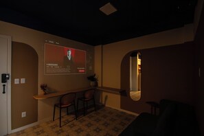 A room with a dark ceiling, a wall-mounted screen displaying a movie poster, a small seating area with two chairs, and a mirror on the wall.