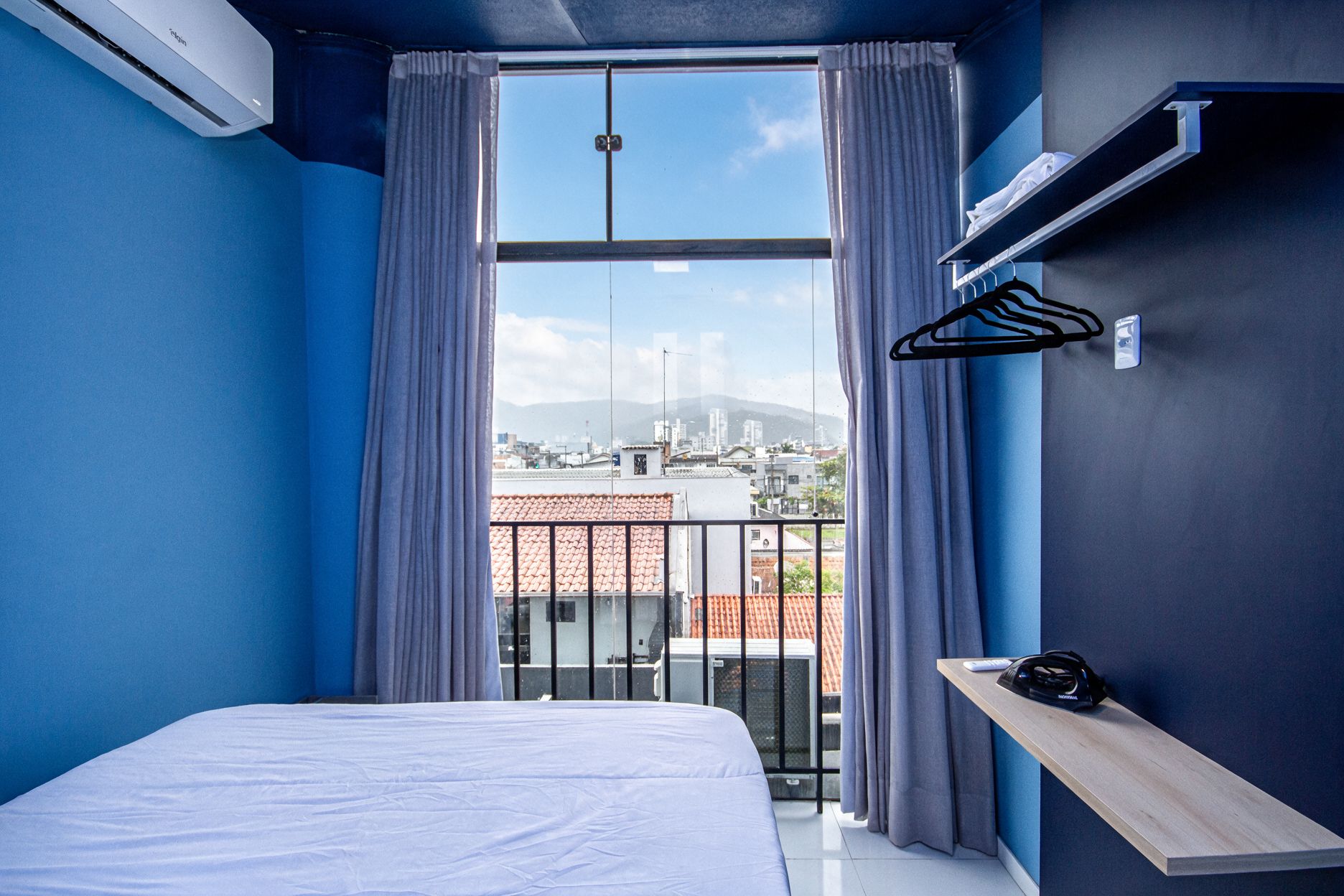 Quarto de hotel com cama, janela com cortinas, vista para a cidade e uma pequena prateleira com controle remoto.