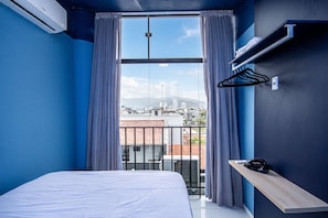 Una habitación de hotel con una cama, una ventana con cortinas, vistas a un paisaje urbano y un estante pequeño con un control remoto.