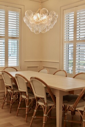 En spisestue med et lyst bord, fletstole, en dekorativ lysekrone og vinduer med hvide rammer.