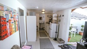 A narrow hallway leading to a kitchen with a refrigerator, sink, and cabinets. There is a door leading to a bathroom, and a small seating area with a chair and a table. - RAS Casita Encanto (West Palm Beach)