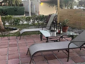 An outdoor patio area with a glass table and metal chairs, surrounded by a brick floor and a wooden fence. - RAS Casita Encanto (West Palm Beach)