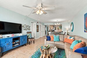 A living room with a television, blue entertainment unit, glass coffee table, and a beige sofa with colorful cushions. - Sand Castle Oceanfront Condo (Indian Shores)