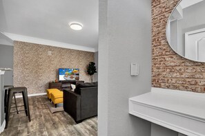 A modern living room with a brick-patterned wall, a flat-screen TV, a dark sofa, and a yellow ottoman. - 2BR Condo Near Disney Resort Pool (Kissimmee)
