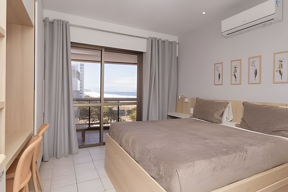 A hotel room with a bed, a desk, a chair, a balcony with a view, and artwork on the wall.