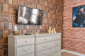 A room with a brick wall, a bookshelf filled with books, a flat-screen TV mounted on the wall, and a dresser with drawers. - 8BR Near Disney Private Pool, spa & Lake View (Kissimmee)