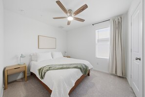 Un dormitorio con cama, mesita de noche, ventilador de techo, ventana con cortinas y puerta.