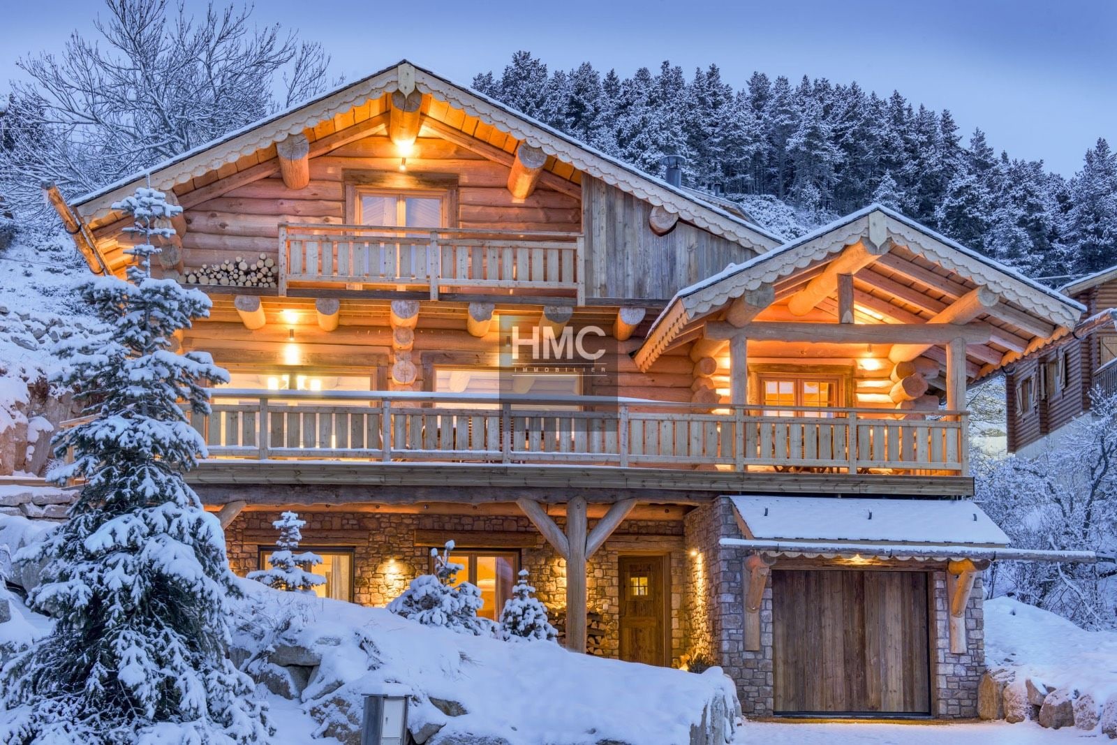 A two-story log cabin with a balcony, stone foundation, and a wooden garage.