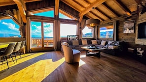 A spacious living area with a wooden floor, a sofa, a coffee table, and a dining table with chairs. Large windows provide a view of the outdoors. - Papa Shelter, vue époustouflante et Bain Nordique (Bolquère)