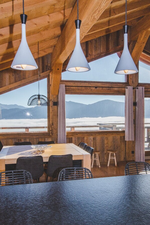 A dining area with a wooden table and chairs, three pendant lights, and a view of mountains through large windows. - Chalet "Nest Lodges" (Les Angles)