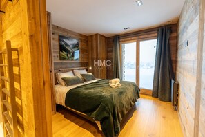 Ein schlichtes Schlafzimmer im Blockhausstil mit einem Bett, Kissen und Blick auf eine verschneite Landschaft durch ein großes Fenster.