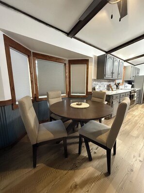 A dining area with a round table and six chairs, a kitchen with grey cabinets, and a window with blinds. - House in Seeley Lake perfect for a relaxing getaway (Seeley Lake)