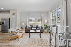 A modern living room with a sofa, coffee table, and chairs, a kitchen area with a refrigerator, and large windows with blinds. - 5BR Townhouse Near Disney Pool & Oasis Club (Davenport)