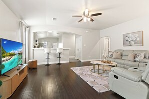 A modern living room with a flat-screen TV, a sofa, a coffee table, and a ceiling fan. - Cozy Modern 3br/2ba Retreat Near 3 Highways (Houston)