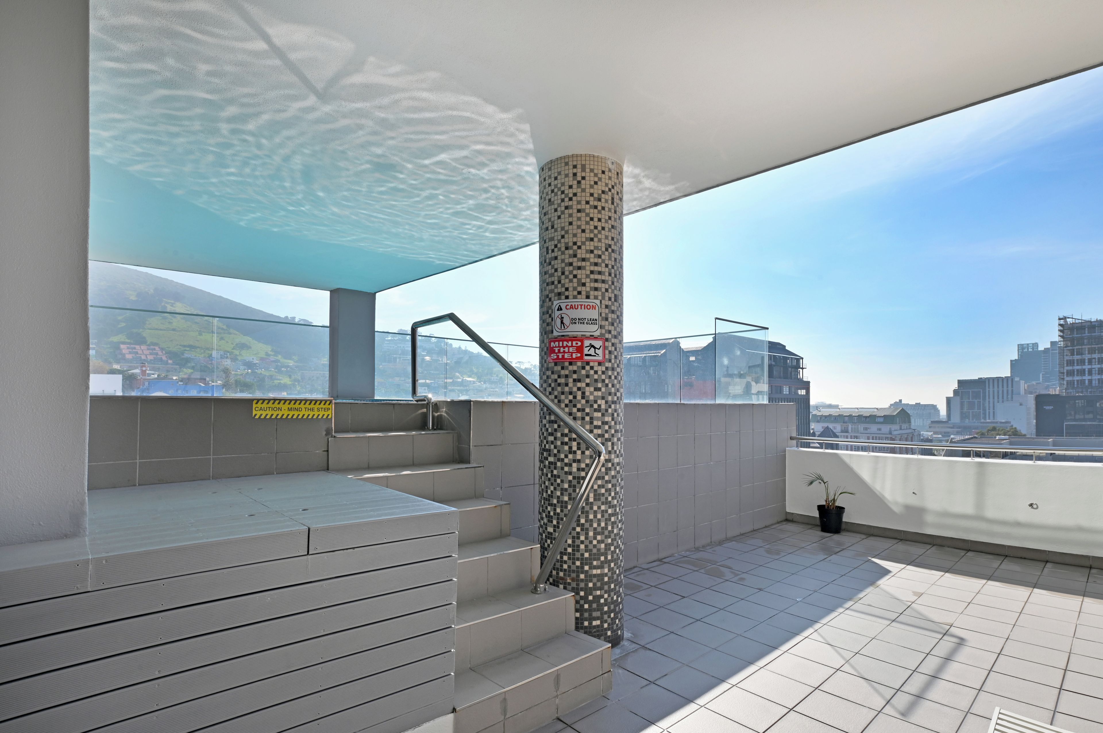 A rooftop terrace with a glass balustrade, a tiled floor, and a view of a cityscape.