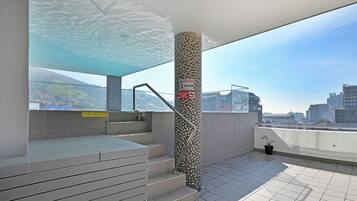 A rooftop terrace with a glass balustrade, a tiled floor, and a view of a cityscape.