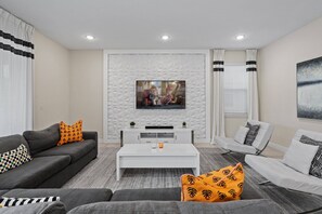 A modern living room with a flat-screen TV mounted on a textured wall, grey sofas with patterned cushions, a white coffee table, and a white armchair. - 6BR Near Disney Private Pool & Game Room (Kissimmee)