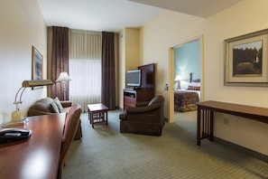 A hotel room with a wooden desk, a brown armchair, a television, a bed with a patterned bedspread, and a framed picture on the wall. - Free Breakfast! 1 Bedroom Suite in Tallahassee (Tallahassee)
