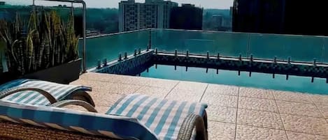 Un espacio al aire libre iluminado por el sol con piscina, sillas de descanso y vistas a edificios lejanos.
