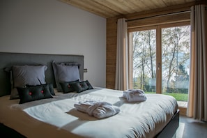 A bedroom with a large bed, grey and black pillows, and a view of trees through the window. - Newly Built Chalet - Charm and Authenticity - Liza's Dacha (Muhlbach-sur-Munster)