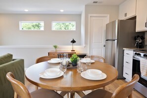 Un comedor con una mesa redonda de madera dispuesta para cuatro personas, rodeada de sillas. Hay una pequeña planta en maceta sobre la mesa, y la habitación tiene un ventanal con vistas a zonas verdes.