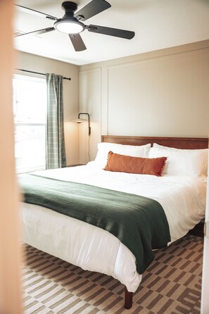 Una habitación de hotel con una cama, ventilador de techo, ventana con cortinas y una lámpara.