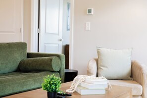 Una sala de estar con un sofá verde, un sofá color beige, una mesita de centro con libros y una planta, y una puerta al fondo.