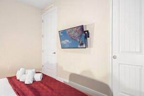 A hotel room with a bed, a red blanket, white towels, a wall-mounted TV displaying a movie scene, and a white door. - 10br Near Disney Private Pool, spa & Game Room (Kissimmee)