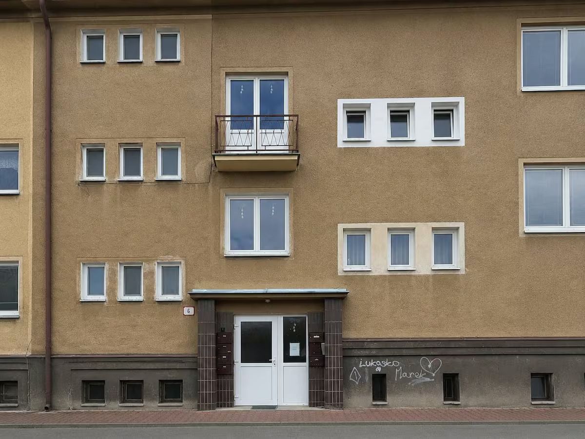Ein mehrstöckiges Gebäude mit einer glatten Fassade, einem zentralen Eingang mit einer Tür und einem kleinen Balkon im zweiten Stock.
