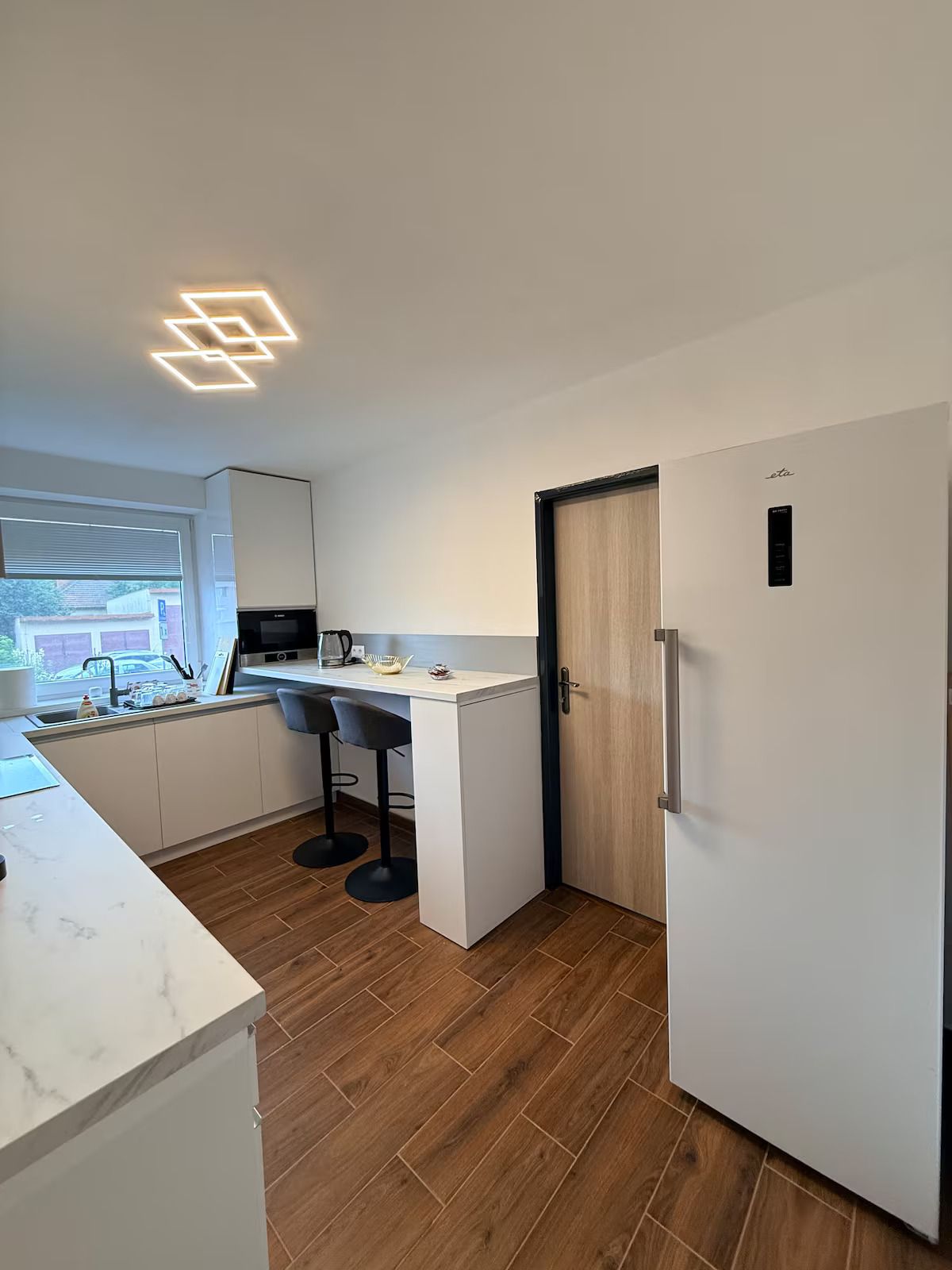 Et moderne køkken med en indbygget ø, et køleskab og et vindue med rullegardiner.