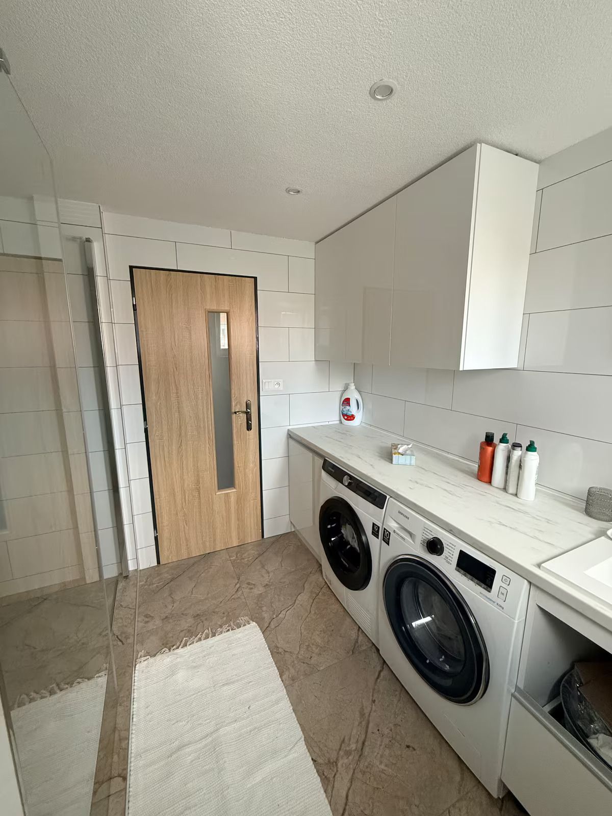A laundry room with a washing machine, a dryer, a wooden door, and a white tiled wall.
