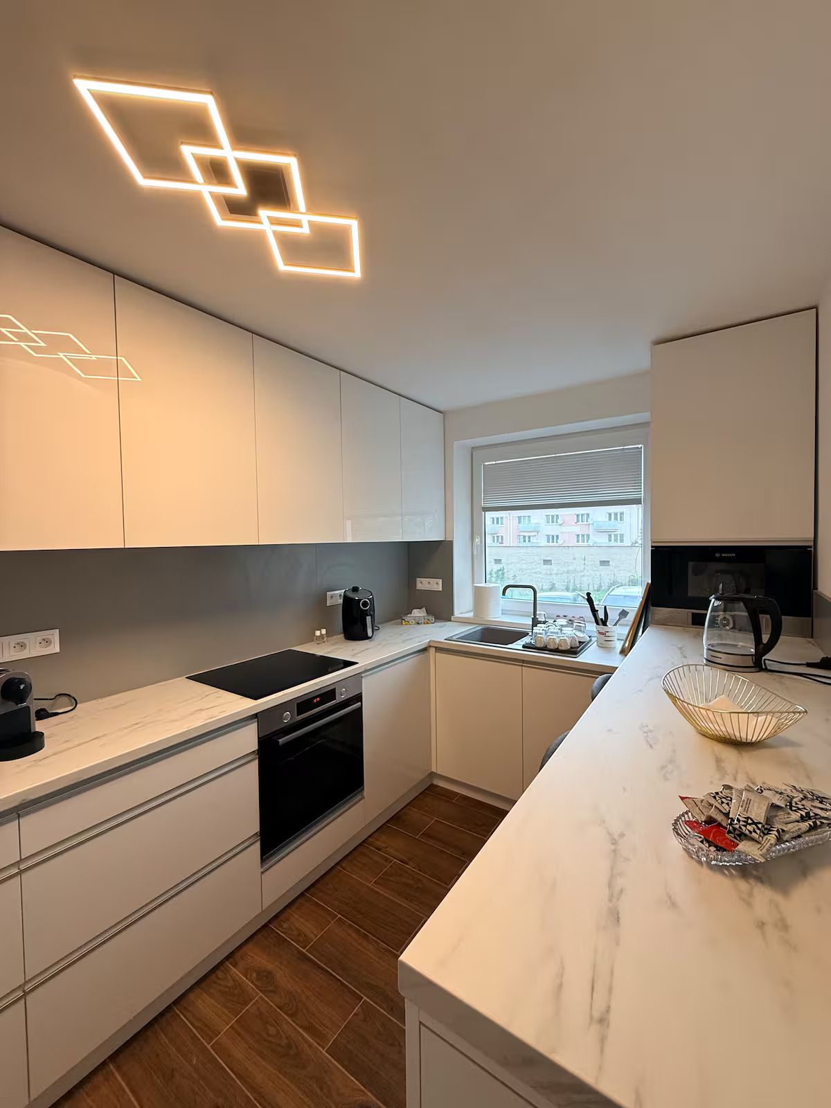 Una cuina moderna amb armaris blancs, forn integrat, cafetera i finestra amb vista a edificis.