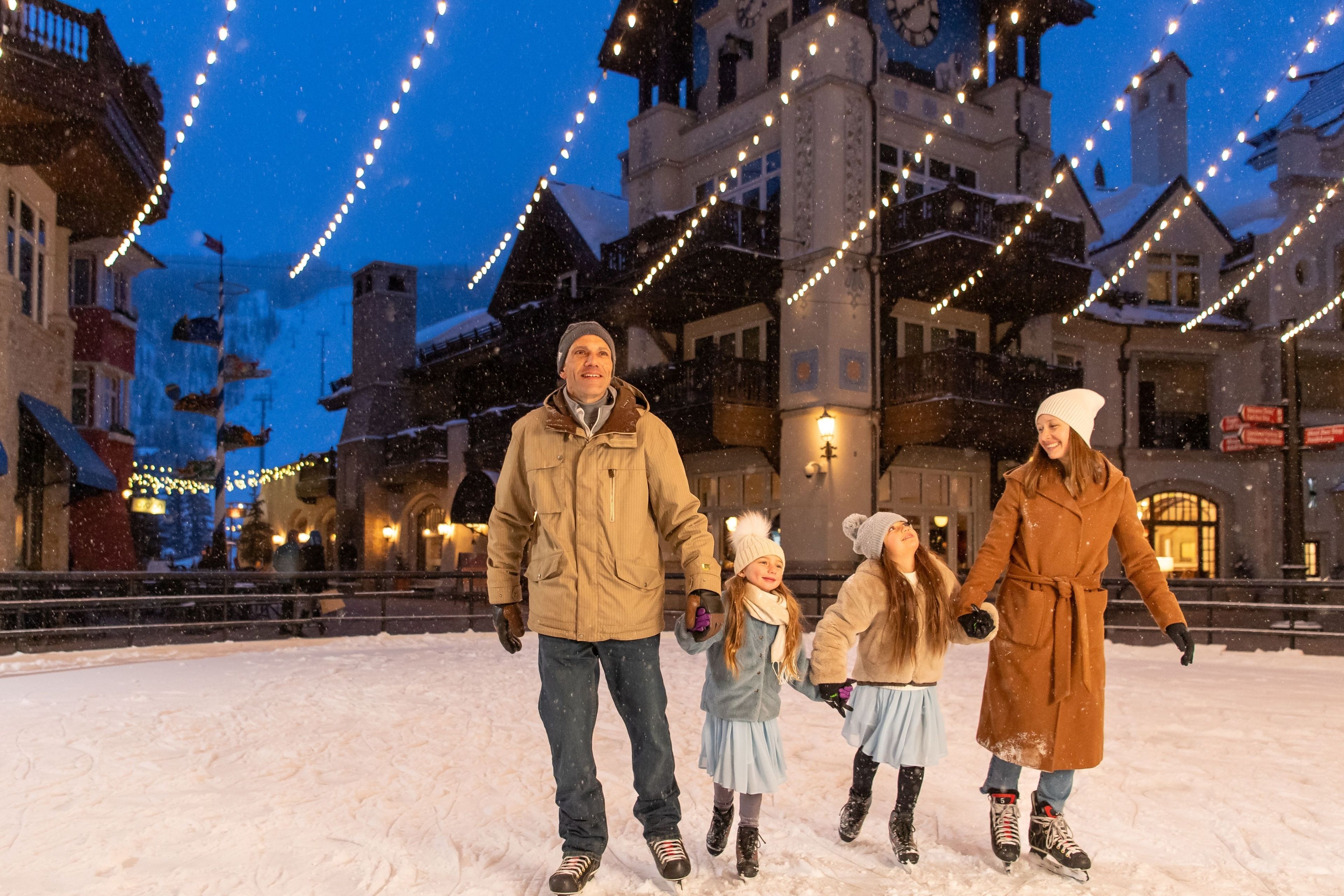 Une famille patine sur une patinoire en plein air, dans un paysage enneigé, avec des bâtiments en arrière-plan.