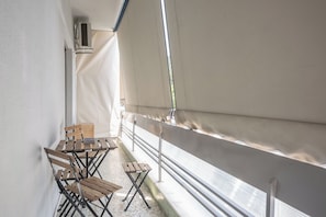 En balkon med et bord og stole, et hvidt gardin og et vindue med persienner.