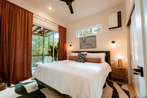 A bedroom with a large bed, a ceiling fan, a window with curtains, a wall-mounted artwork, and a nightstand with a lamp. - Birch & Beard | Boutique Forest Stay + Hot Tub (Young Harris)