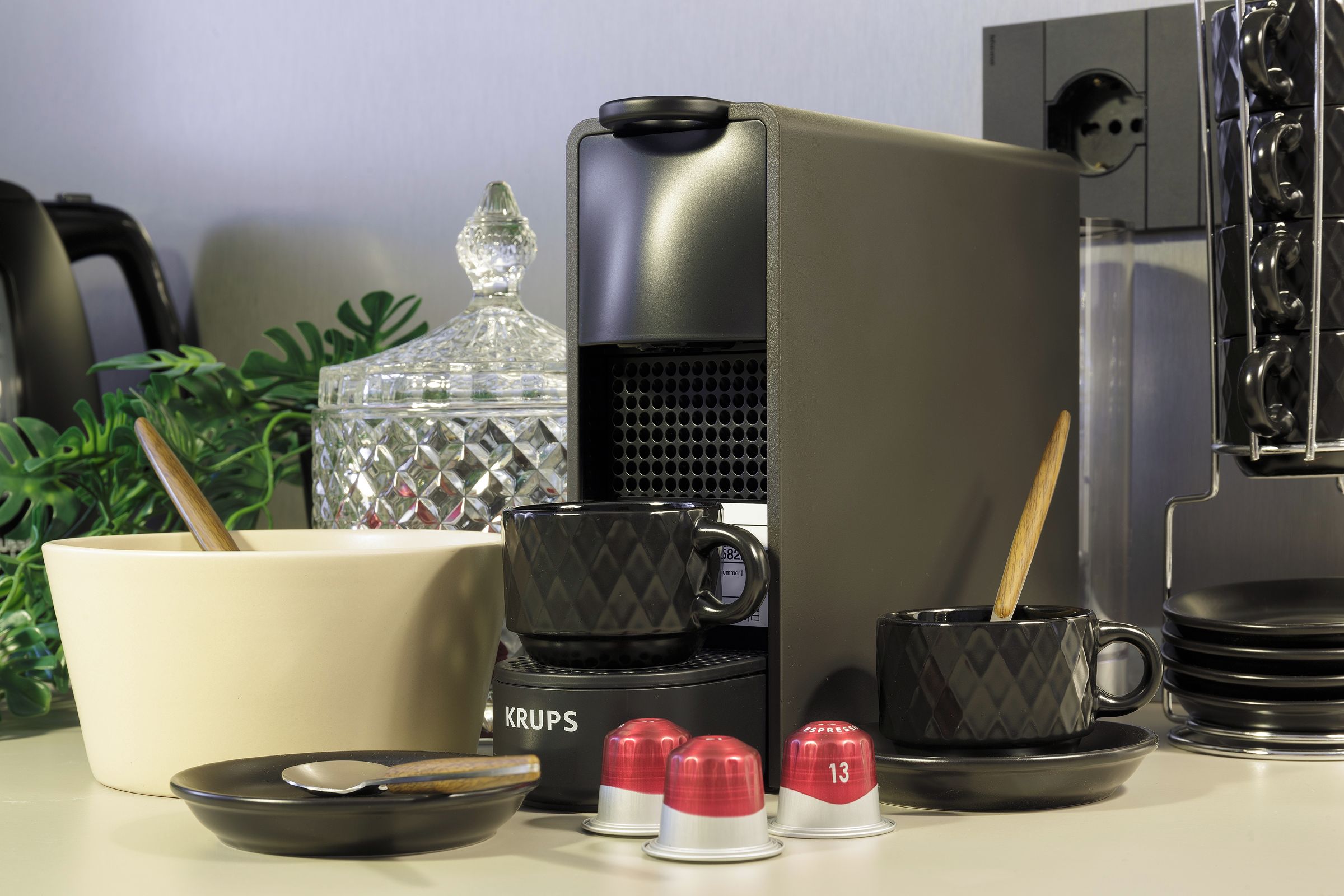 Un área de cocina con cafetera, un recipiente de vidrio, una taza, una cuchara y una taza pequeña con un número.