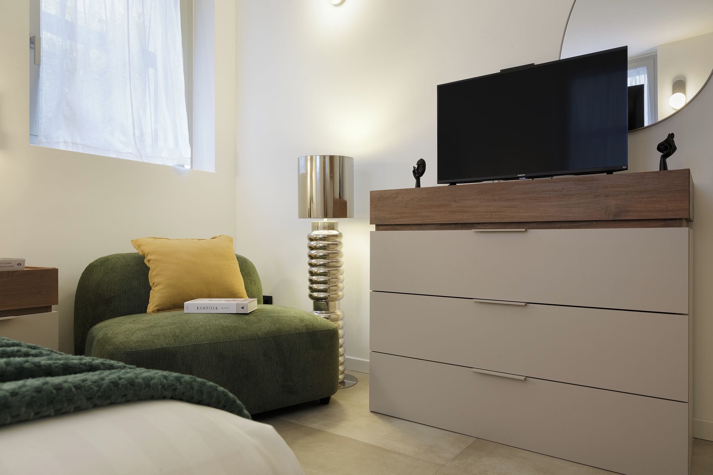 Een moderne slaapkamer met een bed, een stoel, een televisie op een houten dressoir en een lamp.