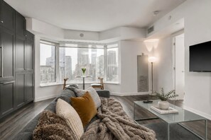 A modern living room with a large window, a sofa, a coffee table, and a flat-screen TV. - Bright 1-Bedroom | Steps from Shops & Transit (Toronto)