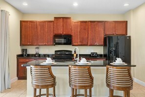 A kitchen with wooden cabinets, a black microwave, a black stove, a black refrigerator, and three wooden bar stools with white plates. - 5BR Near Disney Private Pool, spa & Game Room (Kissimmee)