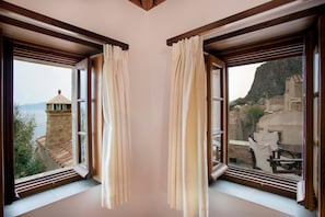 A room with two windows, each with curtains, overlooking a mountainous landscape with traditional architecture. - New Malvasia Hotel (Monemvasia)