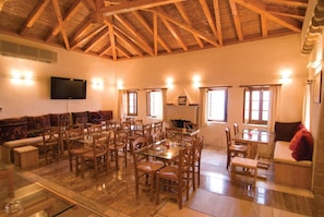 A spacious living area with a high wooden ceiling, a flat-screen TV mounted on the wall, and a dining area with wooden chairs and tables. - New Malvasia Hotel (Monemvasia)