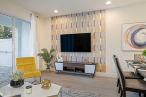A modern living room with a flat-screen TV mounted on a patterned wall, a yellow armchair, a dining area with a table and chairs, and a potted plant. - 3BR Townhouse Near Disney Pool & Mickey Room (Clermont)