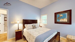 Une chambre à coucher avec un lit en bois, deux tables de chevet, une petite table, une lampe fixée au mur, un tableau encadré et une porte donnant sur une autre pièce.