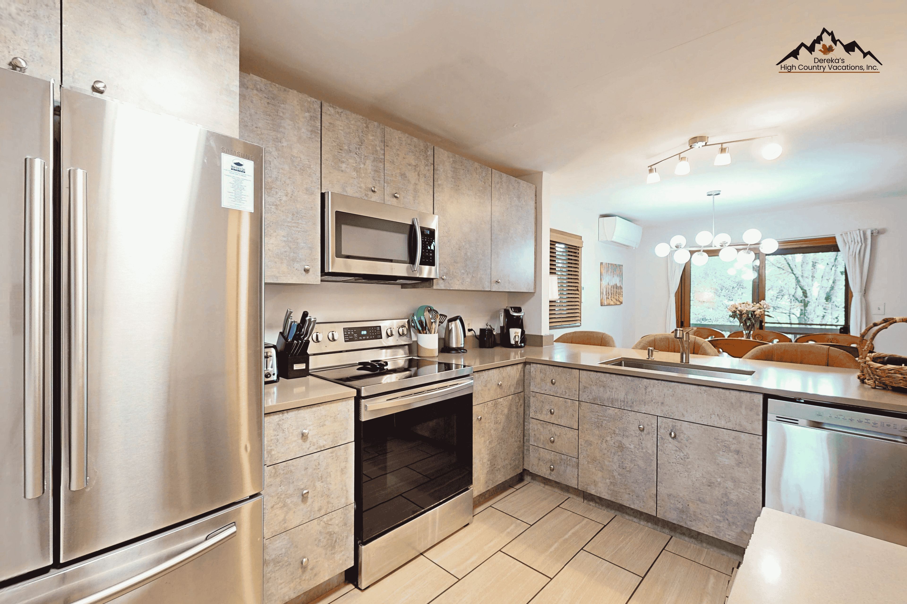 A modern kitchen with stainless steel appliances, a built-in oven, and a microwave. There is a dining area with a window and a chandelier.