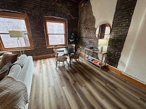 A room with a brick wall, wooden floor, a dining table with chairs, a sofa, and a small table with a lamp. - Historic Brick Loft | Heart of Downtown Knoxville (Knoxville)
