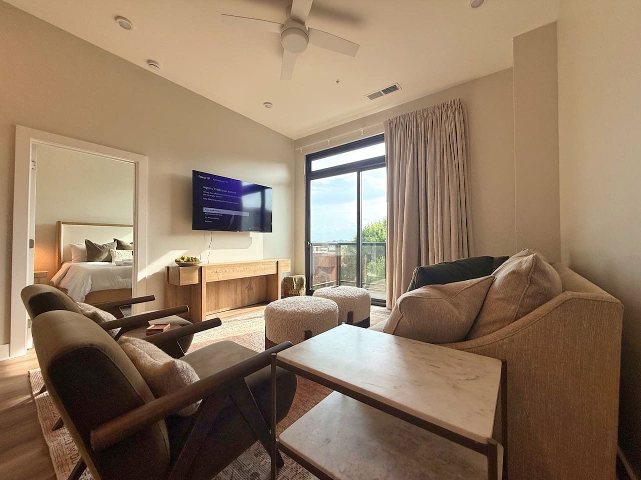 A modern hotel room with a bed, a seating area with a table, a flat-screen TV mounted on the wall, and a large window with curtains.