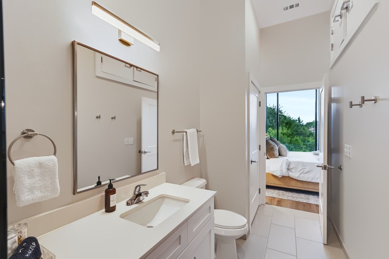A modern bathroom with a large mirror, a sink, and a toilet. There is a towel rack with a white towel and a bottle of soap. The bathroom leads to a bedroom with a bed and a view of greenery outside.