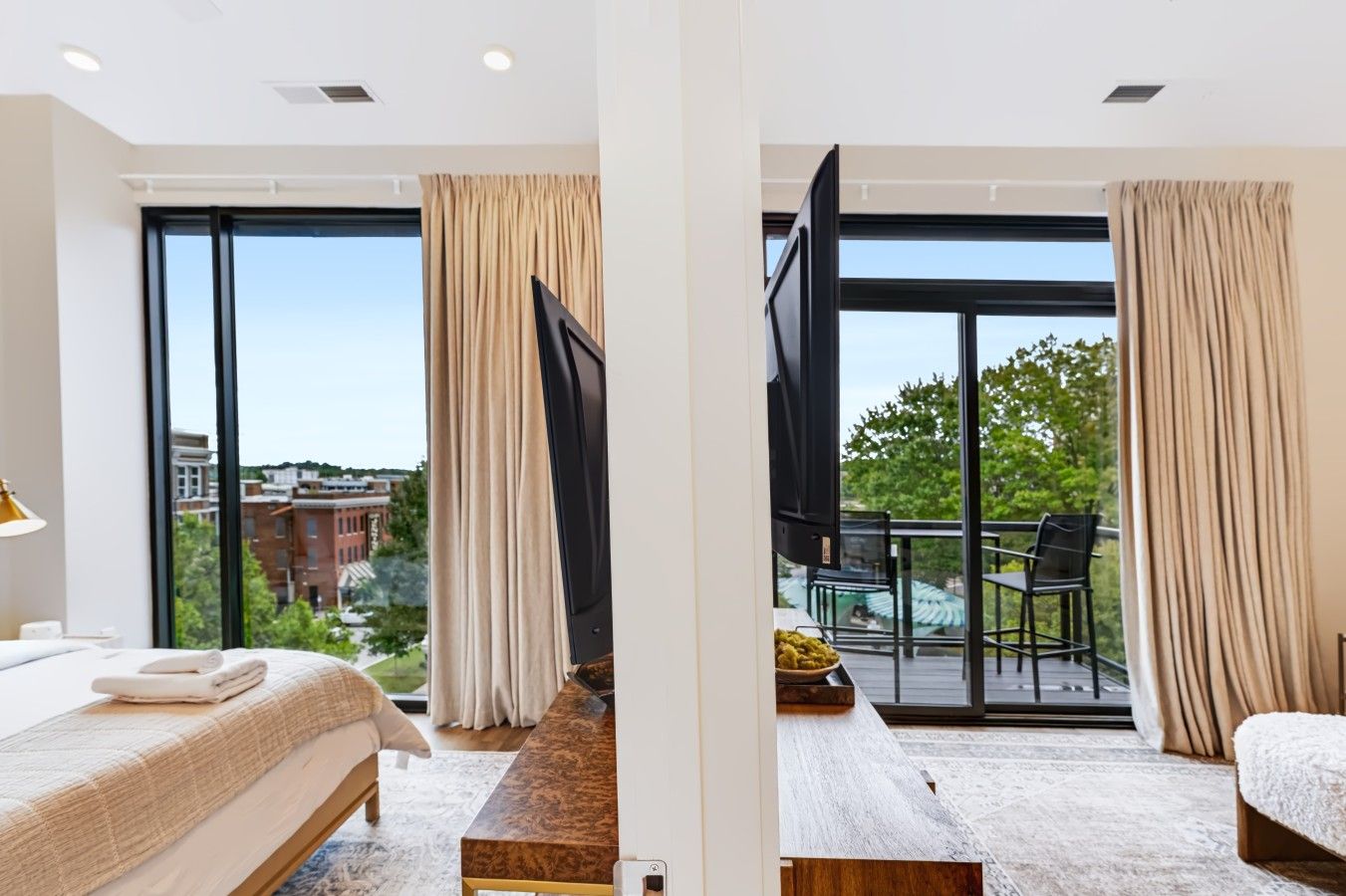 A hotel room with a bed, a TV, a large window with curtains, and a balcony with outdoor furniture.